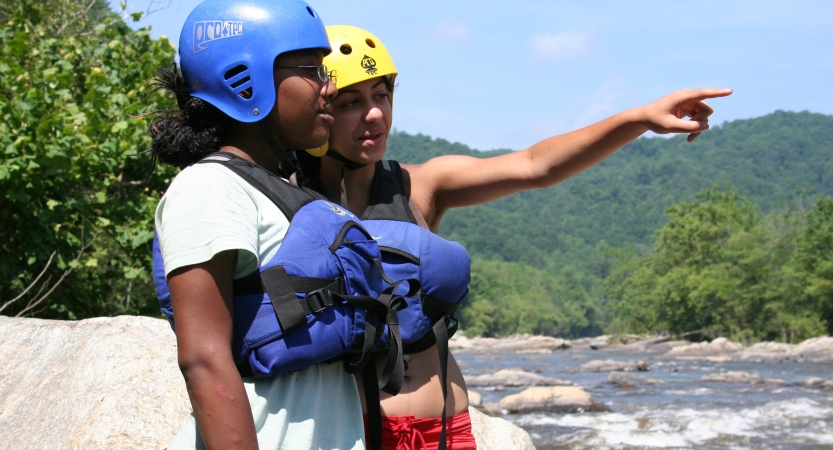 Two people wearing safety gear stand beside a river. One of them points out over the water.